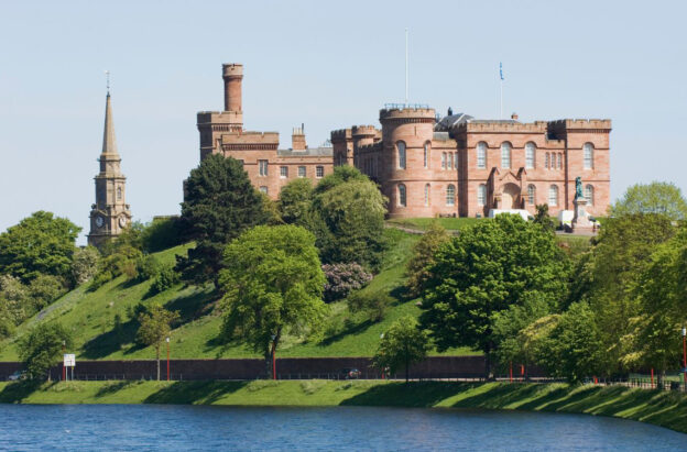 Inverness Castle, red sandstone fortress on a grassy hilltop beside the River Ness, with church spire to the left.